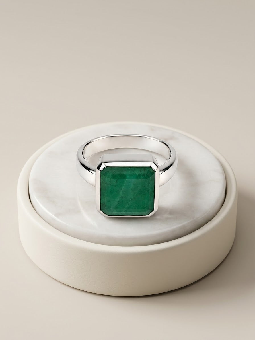 A silver ring with a square-cut green emerald gemstone, displayed on a round white marble and cream ceramic stand against a warm beige background.