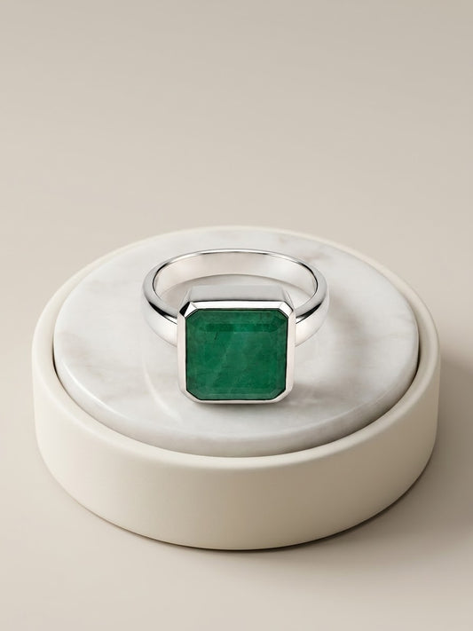 A silver ring with a square-cut green emerald gemstone, displayed on a round white marble and cream ceramic stand against a warm beige background.