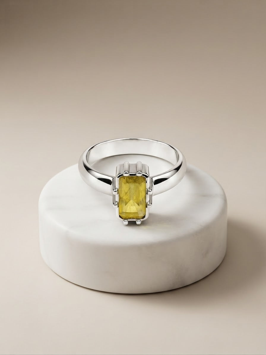 A silver ring with a rectangular yellow gemstone, displayed on a round white marble pedestal against a warm beige background.