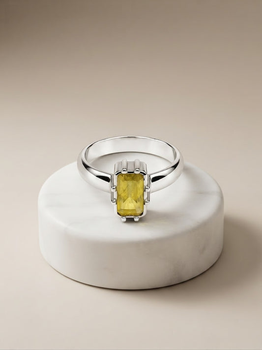 A silver ring with a rectangular yellow gemstone, displayed on a round white marble pedestal against a warm beige background.