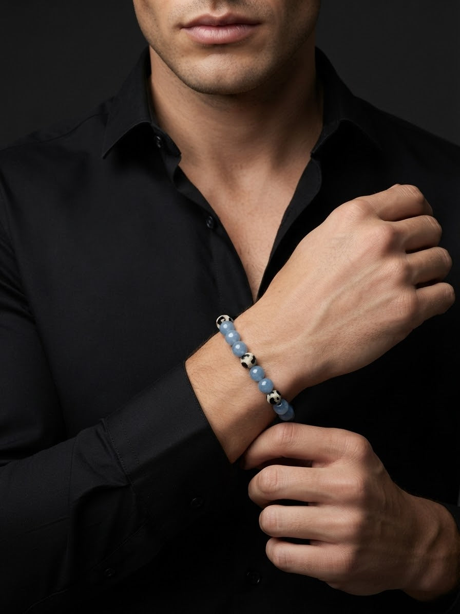 A close-up of a man wearing a beaded bracelet on his wrist, dressed in a black button-down shirt against a dark background.