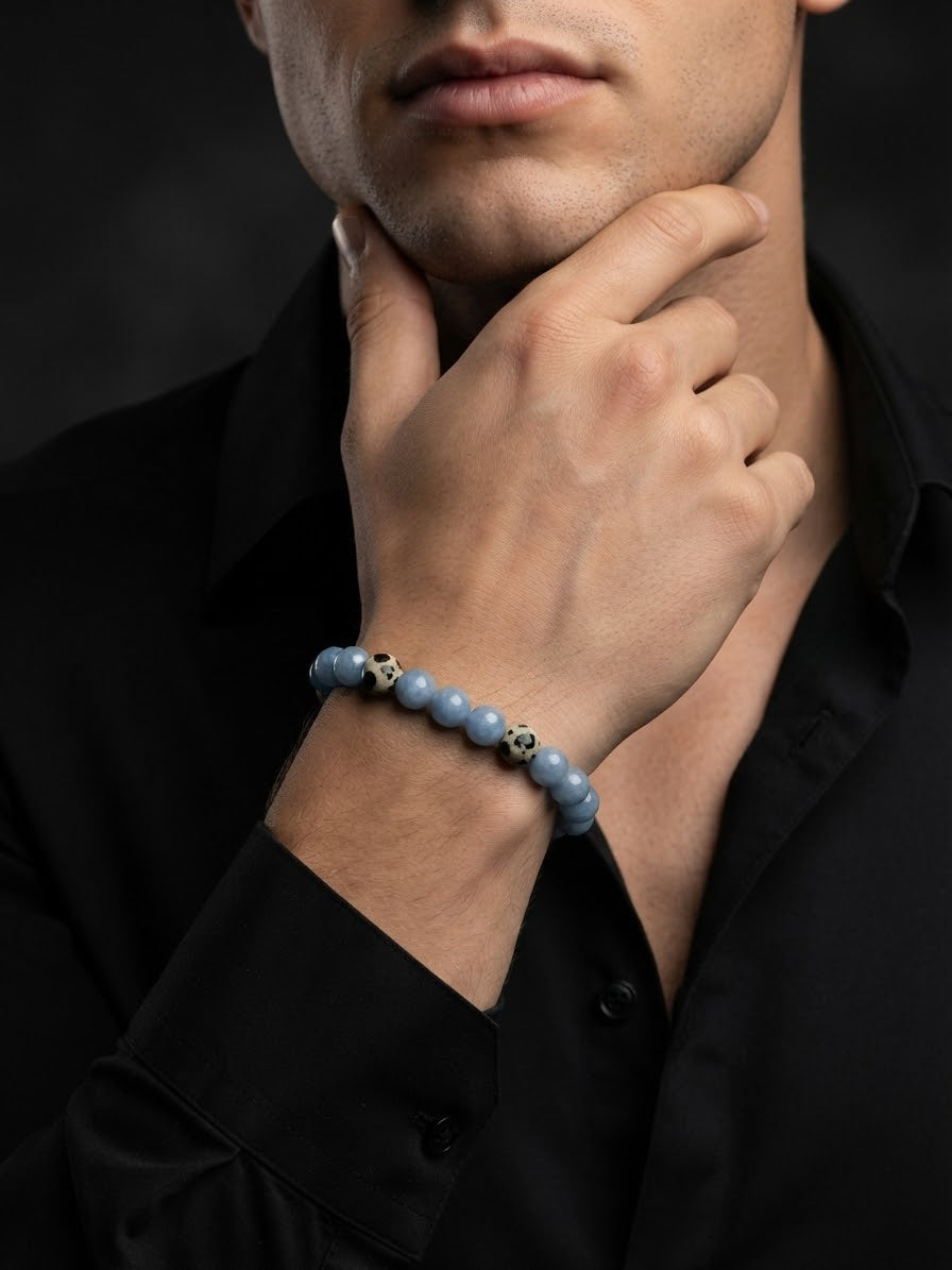 A close-up of a man wearing a light blue beaded bracelet with small gold and silver skull accents, paired with a black shirt against a dark background.