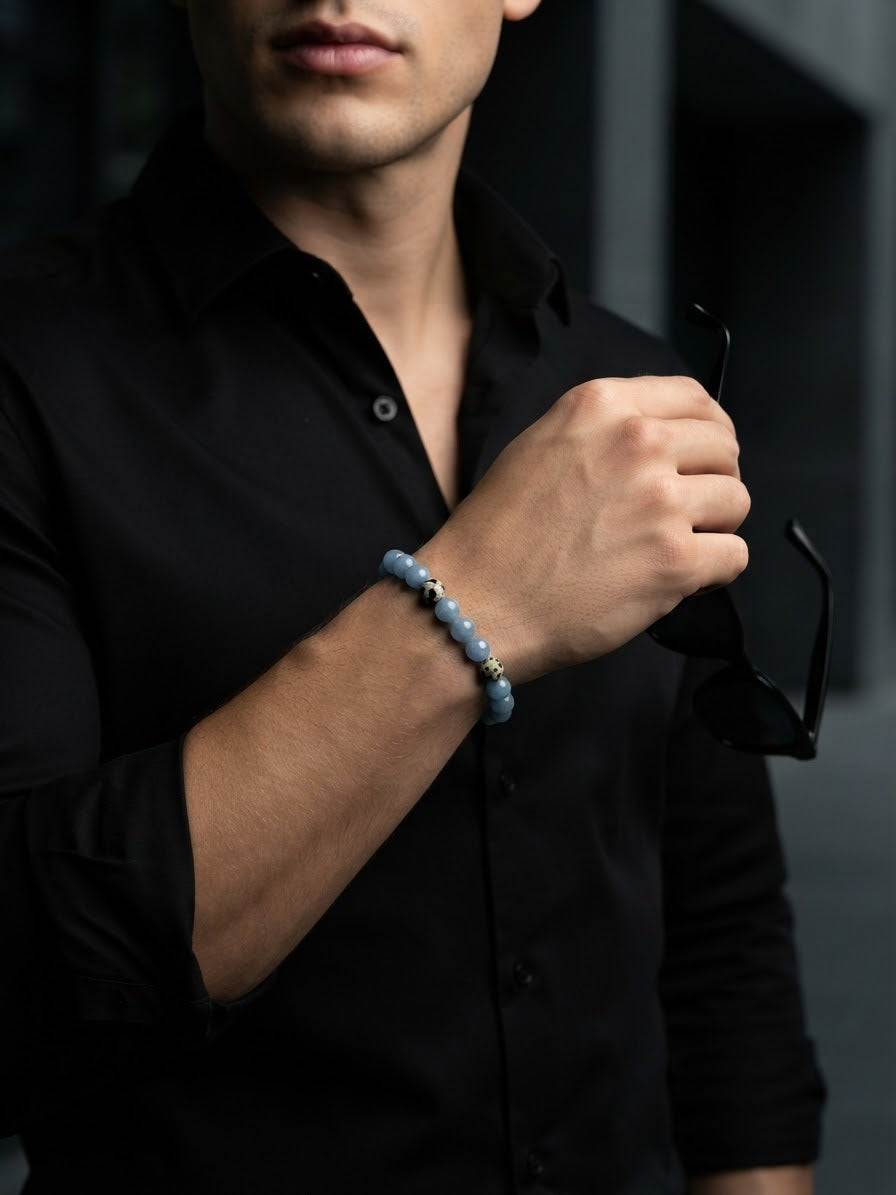 A man wearing a black button-down shirt and holding black sunglasses while wearing a light blue beaded bracelet on his wrist.