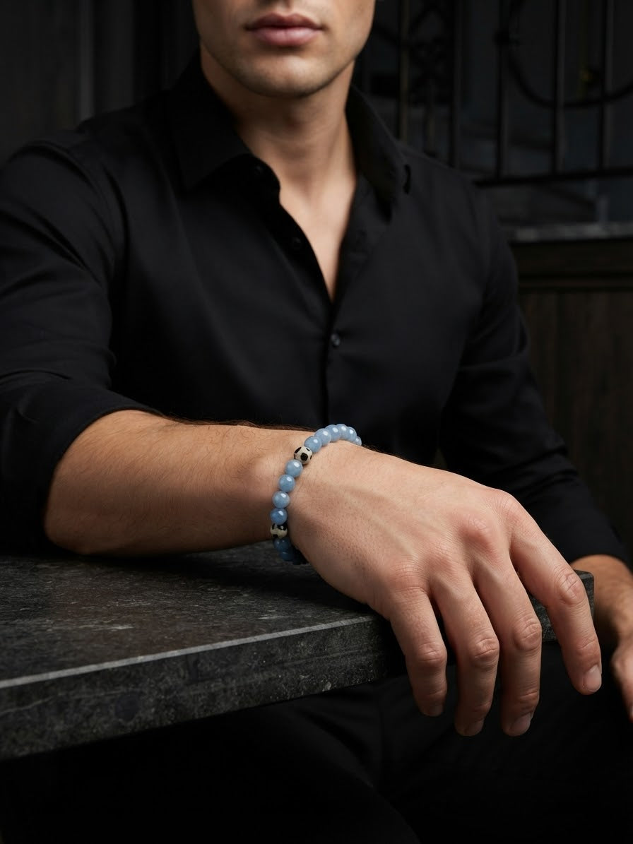 A man wearing a black button-down shirt rests his arm on a dark stone surface, showcasing a light blue beaded bracelet with a small gold accent on his wrist.
