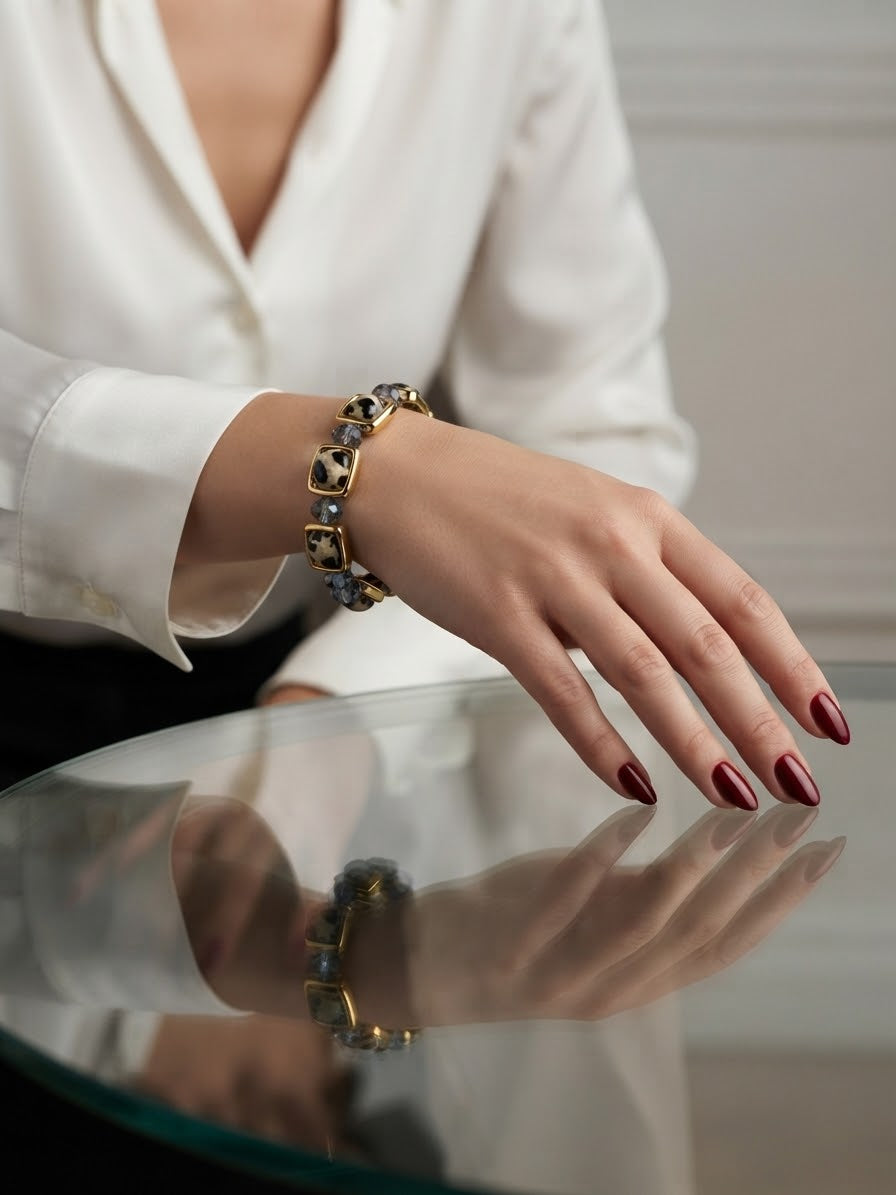 A close-up of a person's hand wearing a gold and dark gray beaded bracelet, resting on a reflective glass table while wearing a white blouse.