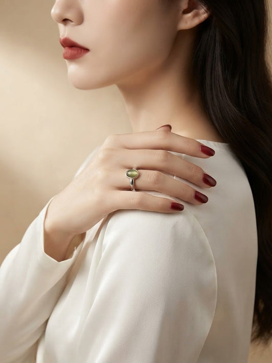 A close-up lifestyle shot of a woman wearing a silver ring with a small oval green stone on her ring finger, dressed in a white satin blouse against a warm beige background.