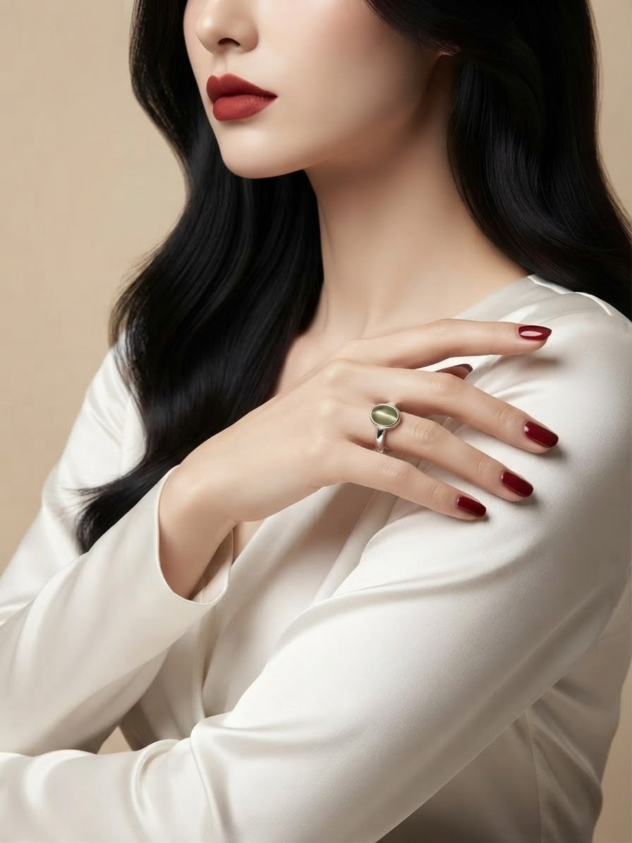 A close-up of a woman wearing a silver ring with a green oval stone, styled with dark red nail polish and a cream satin blouse against a warm beige background.