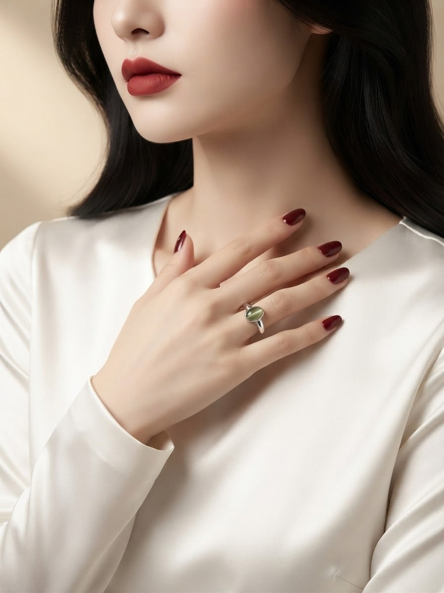 A close-up lifestyle shot of a woman wearing a silver ring with a green marquise-cut gemstone on her finger, paired with a white satin top and dark red nail polish.