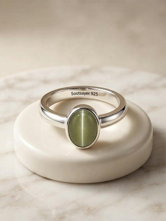 A sterling silver band ring with an oval green cat's eye stone, displayed on a round cream-colored ceramic stand. The inner band is engraved with 'Soostayer 925'.