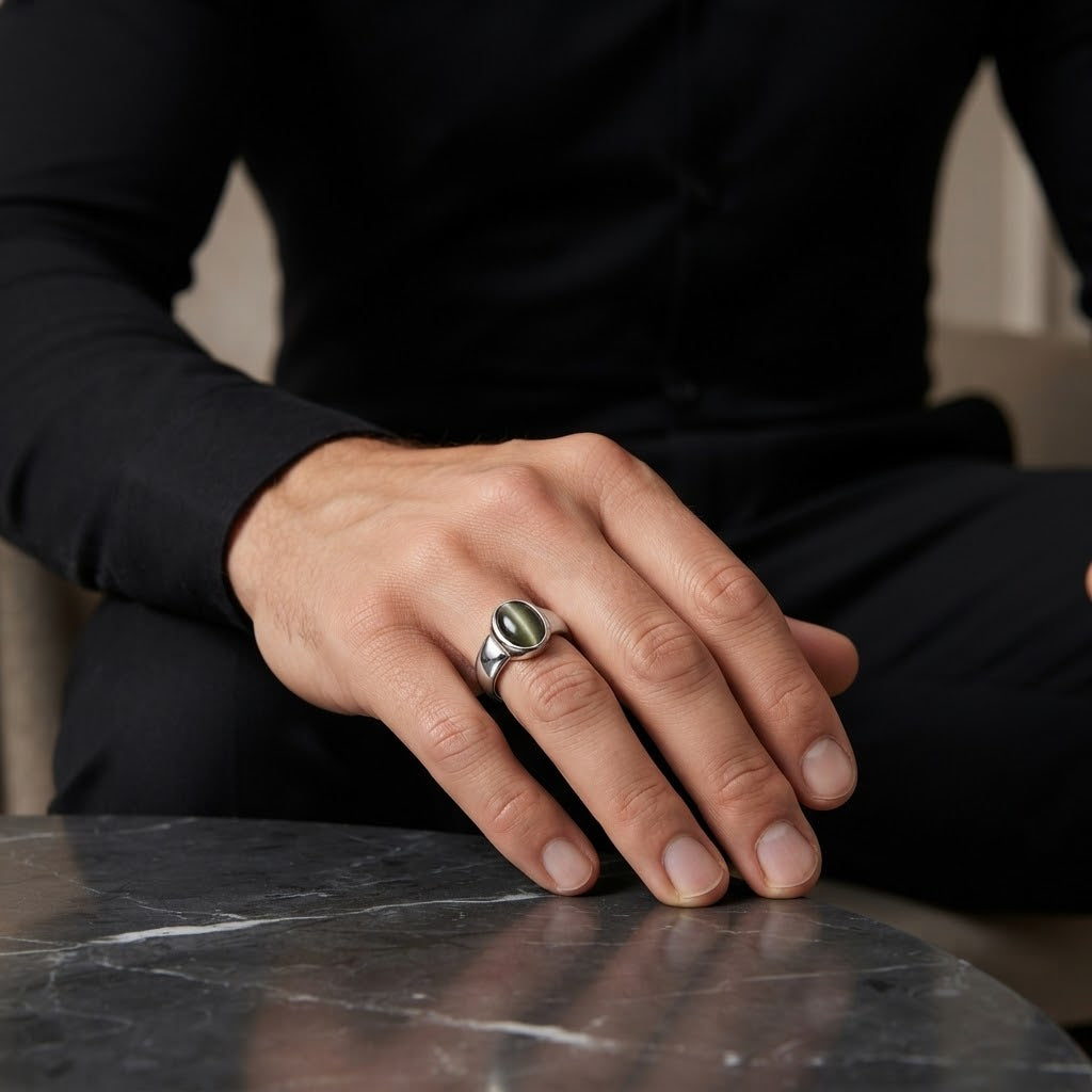 A silver signet ring with a green oval stone worn on a man's hand, which is resting on a dark marble table. The man is wearing a black long-sleeve shirt.