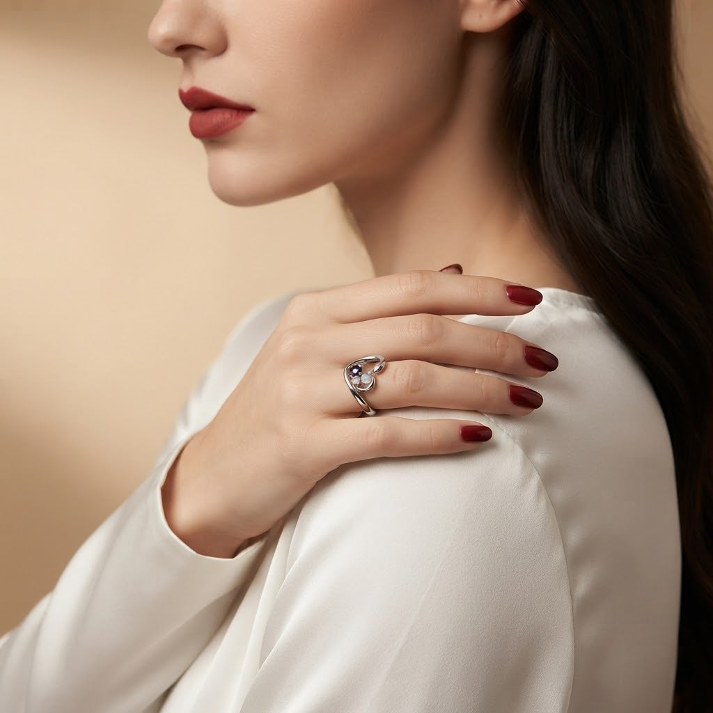 A close-up lifestyle shot of a woman wearing a silver ring with a blue gemstone on her finger, resting her hand on her shoulder. She is wearing a white satin top against a soft beige background.