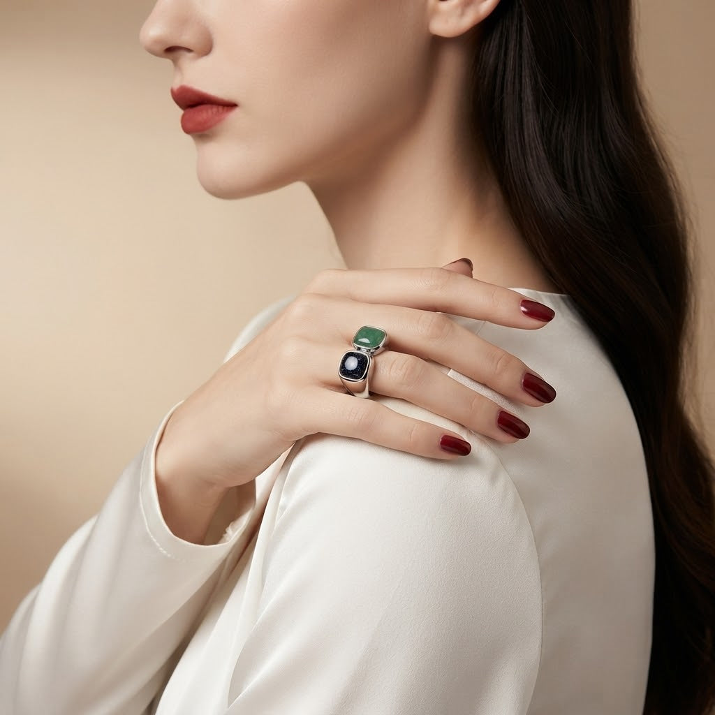 A close-up lifestyle shot of a woman wearing a silver ring with a green rectangular stone and a dark oval stone, shown on her hand against a warm beige background.