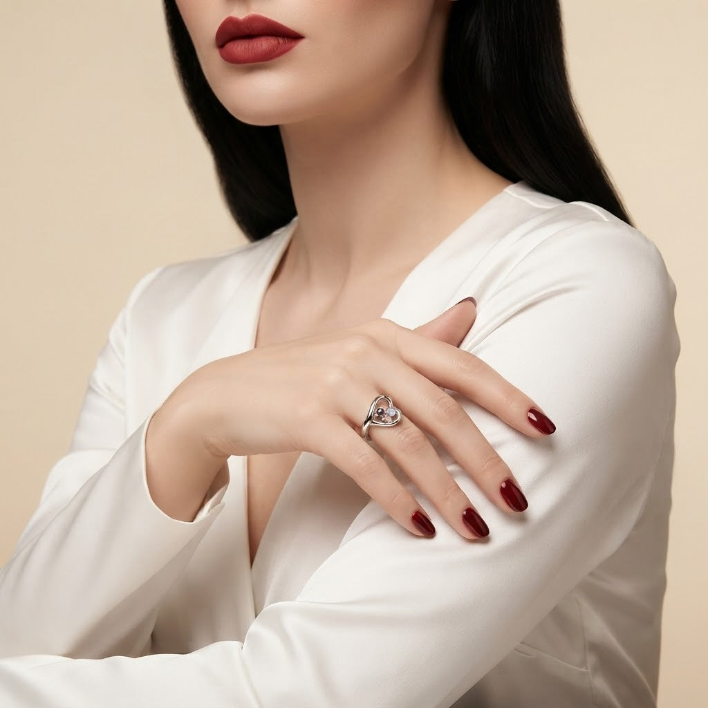 A close-up of a woman wearing a silver ring with a heart-shaped diamond on her finger, styled with a white satin blouse and dark red lipstick against a warm beige background.