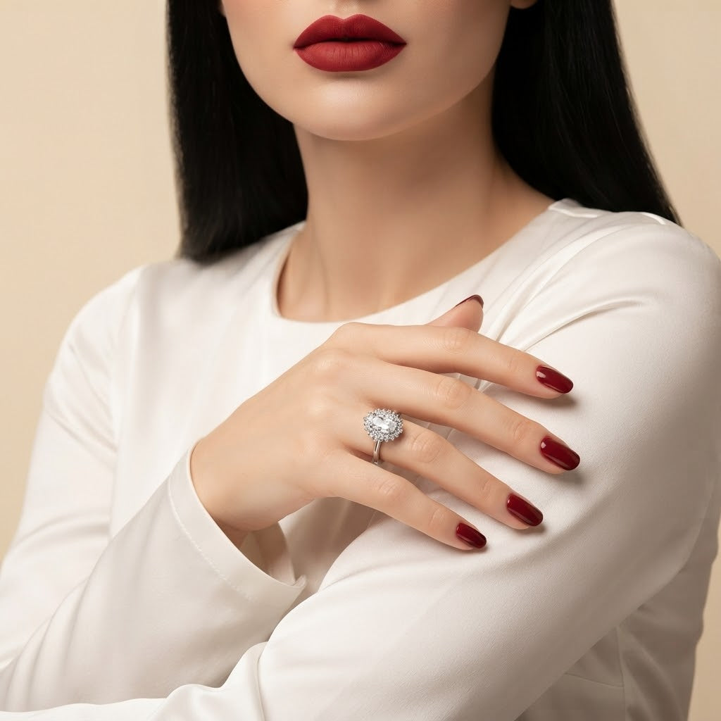 A close-up of a woman wearing a silver-toned diamond engagement ring on her finger, with dark red nail polish and a white long-sleeve top against a warm beige background.