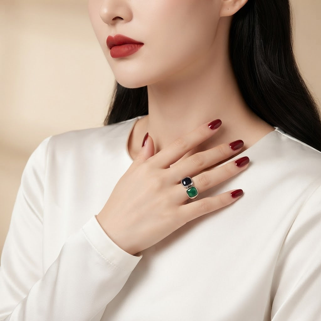 A close-up of a woman wearing a silver ring with a double-stone design, one stone in dark green and the other in black, while wearing a white satin top.