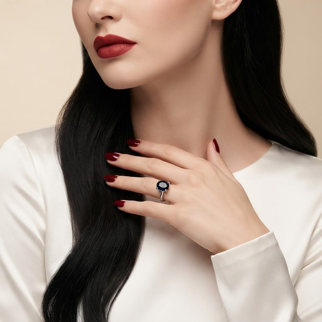 A close-up of a woman wearing a silver ring with a round black gemstone, with her hand resting on her hip. She is wearing a white satin top and has long, straight black hair.