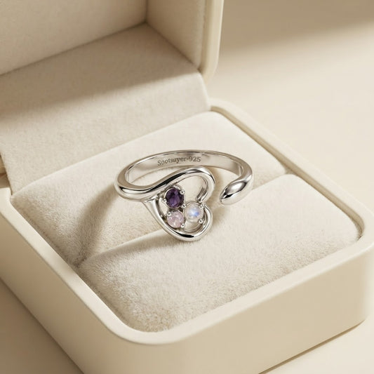 A silver heart-shaped ring with three small colored gemstones (purple, pink, and clear) displayed inside an open cream-colored jewelry box.