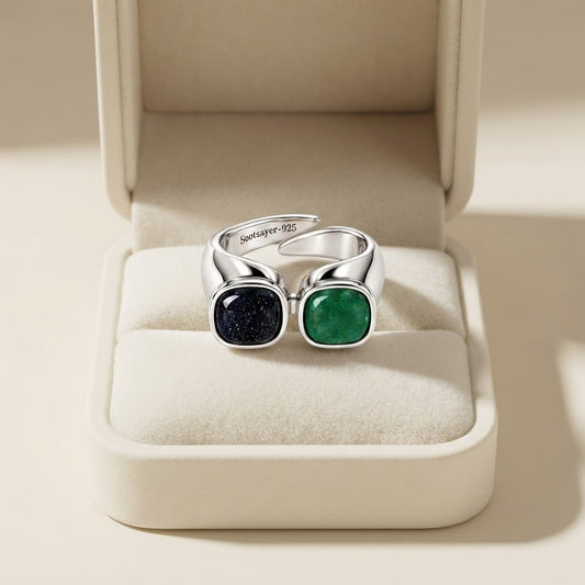 A silver ring with two square bezel-set stones, one dark navy blue and one emerald green, displayed inside an open cream-colored jewelry box.