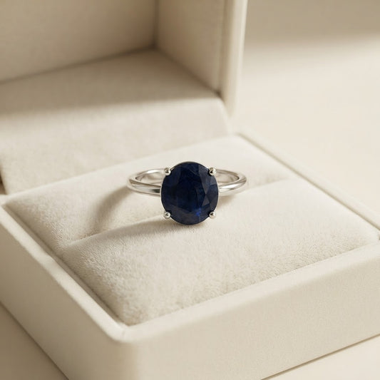 A silver ring with an oval-cut dark blue gemstone resting inside a cream-colored jewelry box.