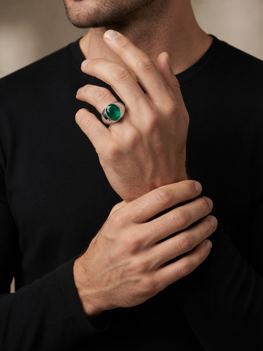 A close-up of a man wearing a silver ring with a round green stone, shown on his hand against a black shirt.
