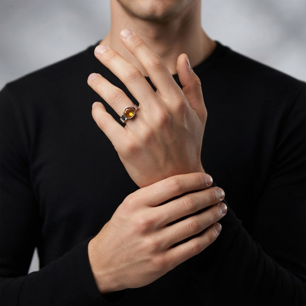 A close-up of a person wearing a ring with a round amber-yellow stone on a dark band, with the hands crossed over the chest against a soft gray background.