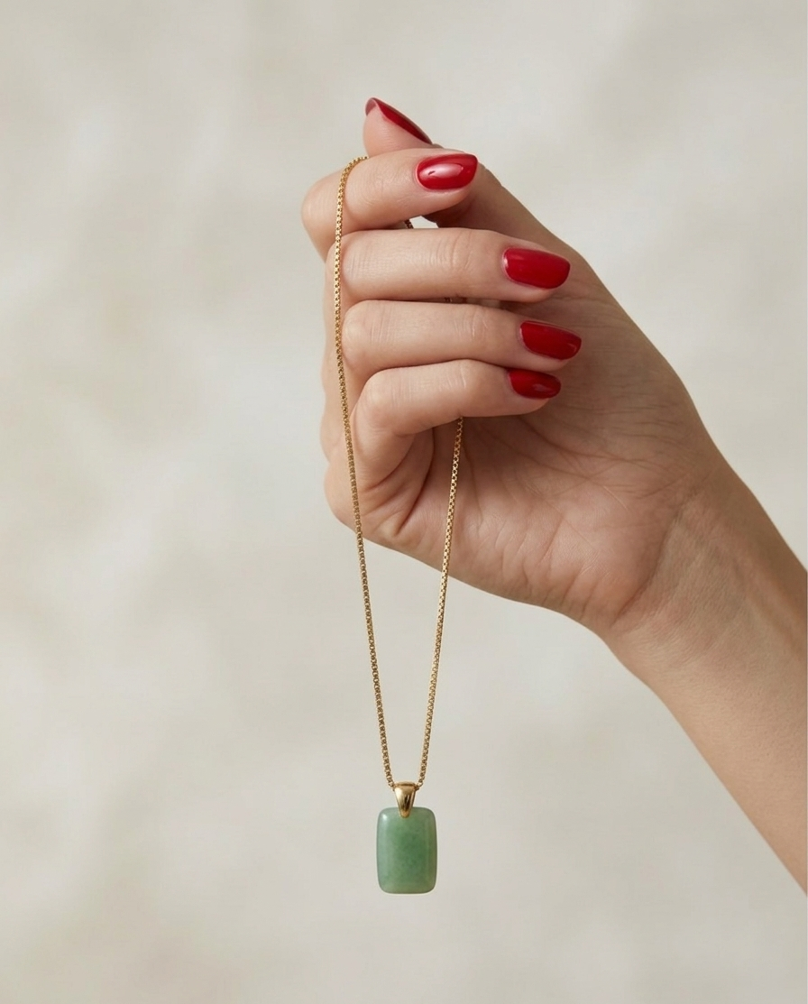 Hand holding a necklace with a green pendant against a neutral background