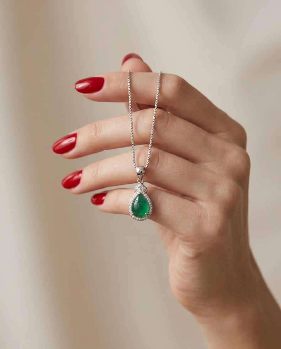 Hand holding a necklace with a green teardrop pendant against a neutral background