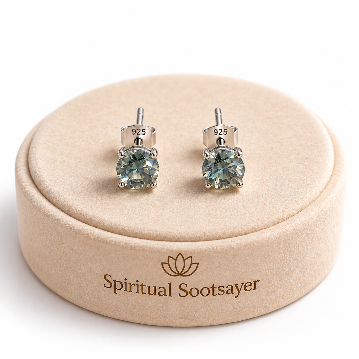 A pair of stud earrings with round faceted light blue-green gemstones in silver settings, displayed on a round beige velvet box engraved with the brand name 'Spiritual Sootsayer'.