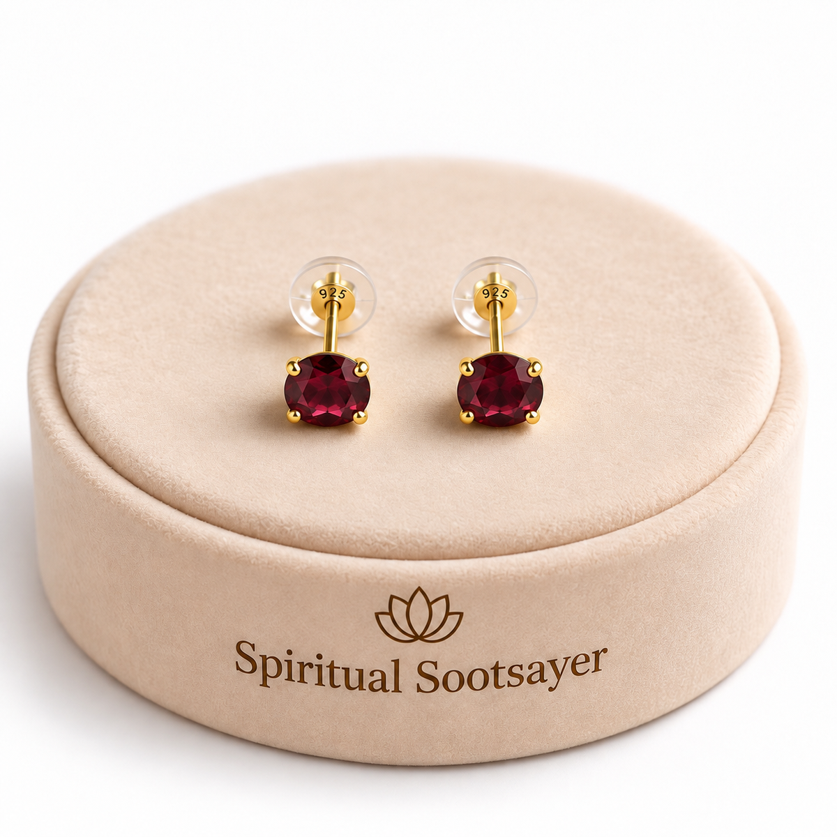 A pair of gold stud earrings with round deep red gemstones, displayed on a beige round jewelry box lid printed with the brand name 'Spiritual Sootsayer'.