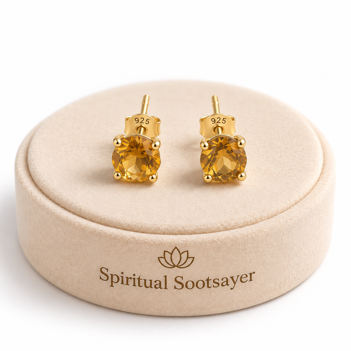 A pair of gold stud earrings with round yellow gemstones, displayed on a round beige jewelry box lid. The box lid is branded with the name 'Spiritual Sootsayer' and has '925' sterling silver stamped on the inside of one earring.