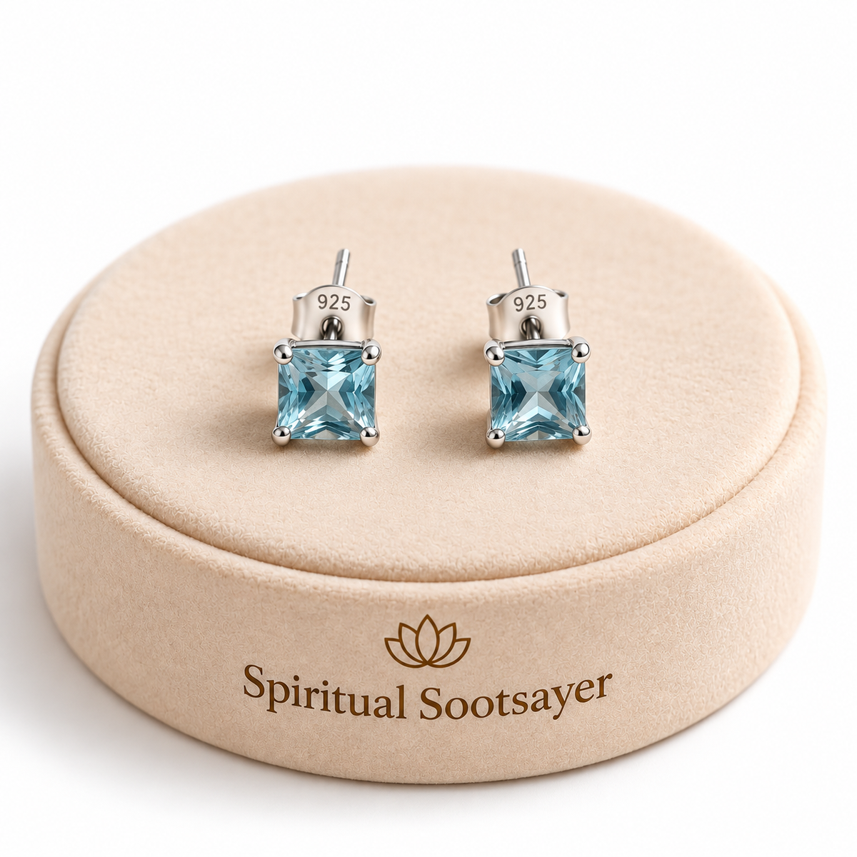 A pair of square-cut light blue gemstone stud earrings in silver settings, displayed on a round beige jewelry box with the brand name 'Spiritual Sootsayer' printed on it.