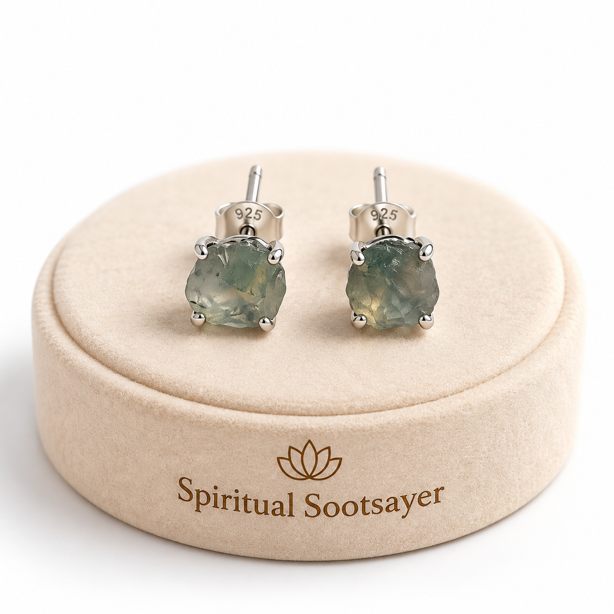 A pair of silver stud earrings with green gemstone settings, displayed on a beige round jewelry box branded 'Spiritual Sootsayer'.