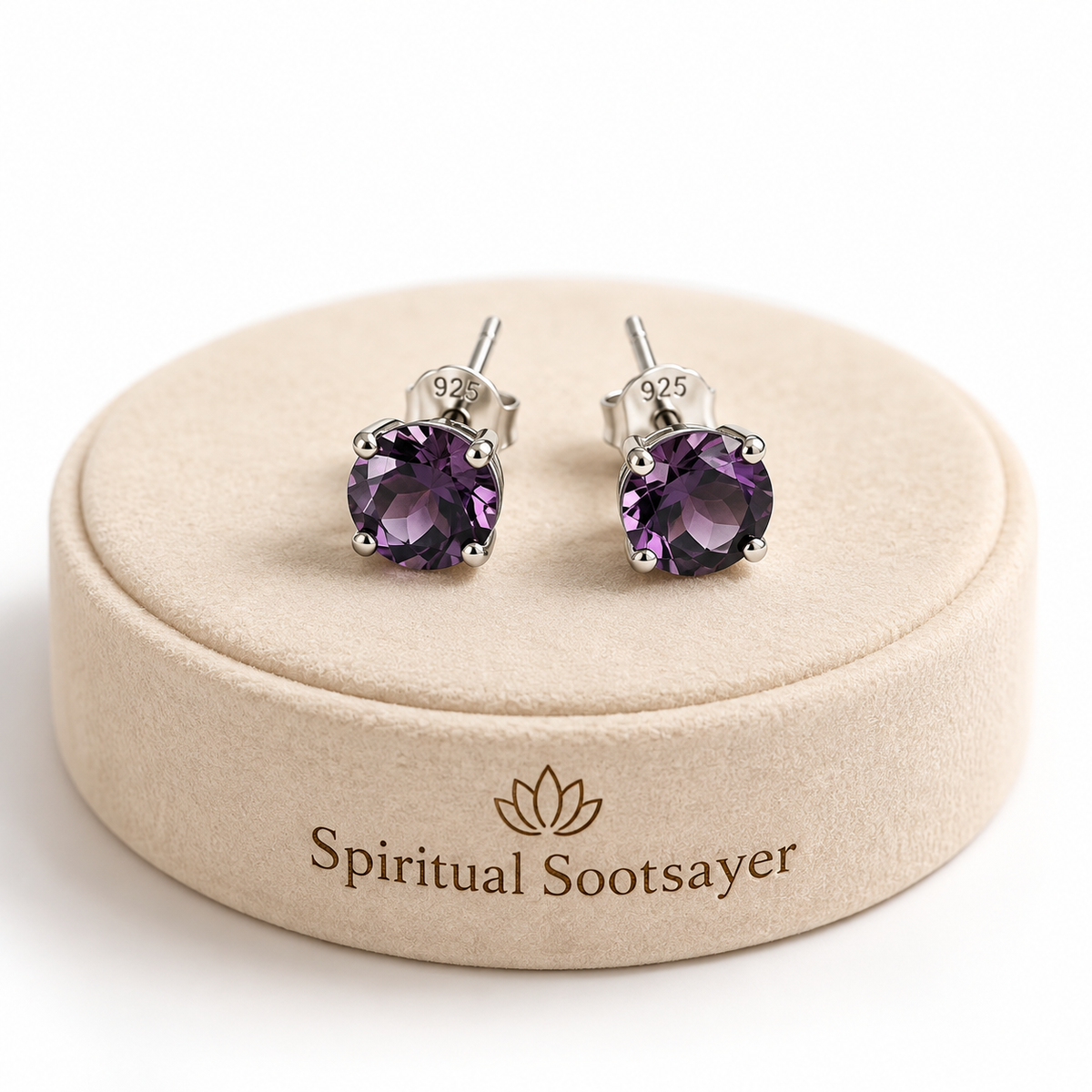 A pair of stud earrings with round purple gemstones in silver settings, displayed on a beige round jewelry box lid printed with the brand name 'Spiritual Sootsayer'.
