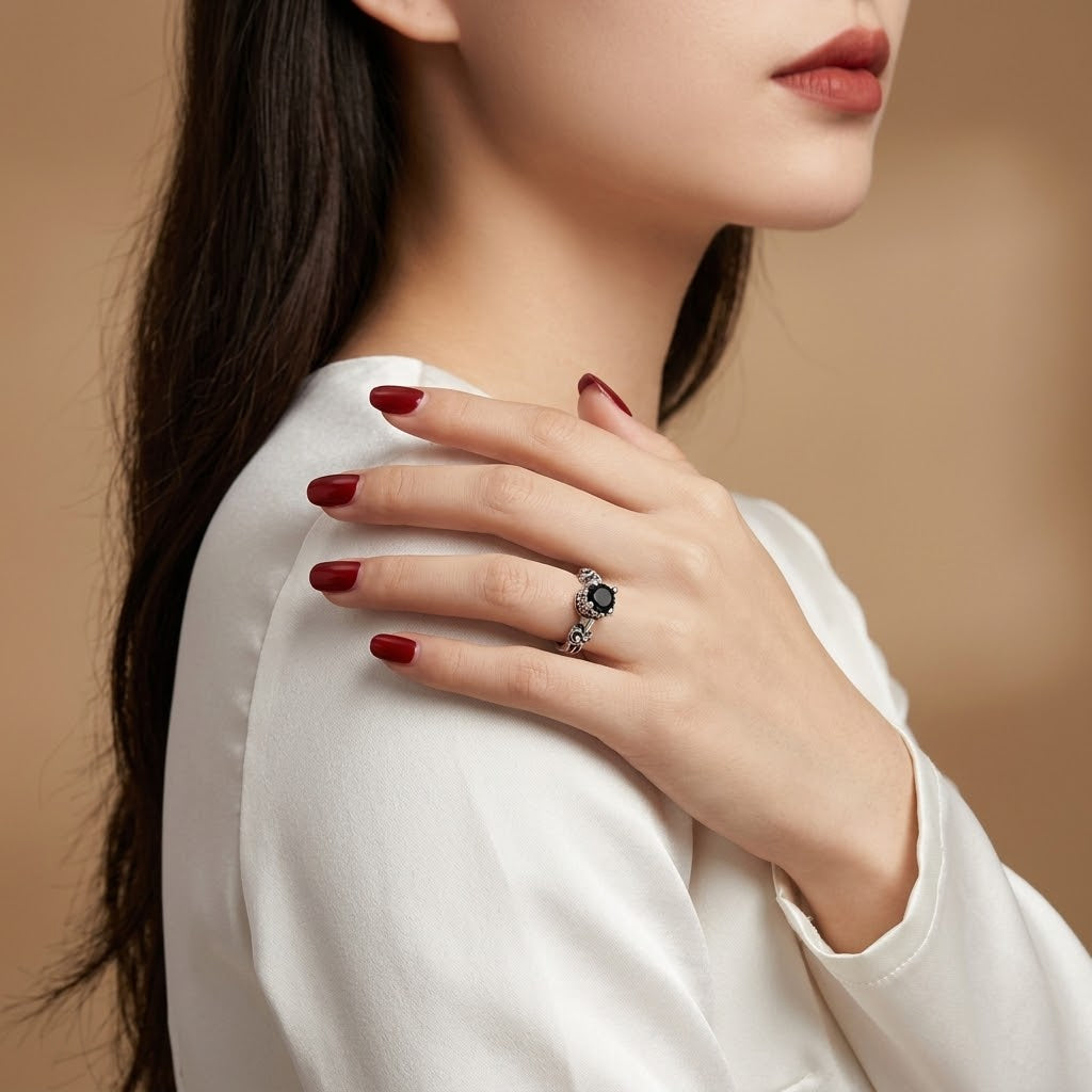 A close-up lifestyle shot of a woman wearing a silver ring with a dark oval gemstone on her hand, which is resting on her shoulder. She is wearing a white long-sleeve top against a warm beige background.