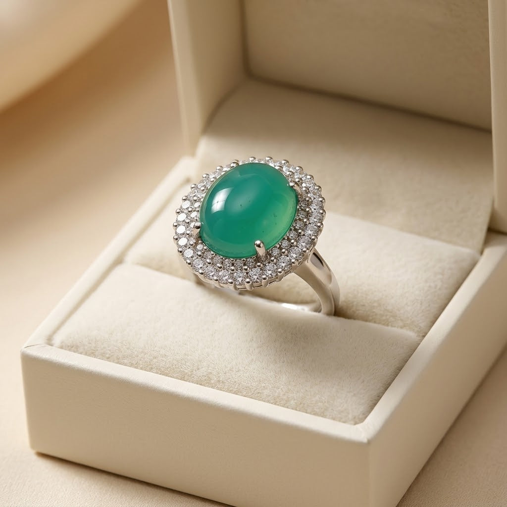 A silver ring featuring a round green cabochon gemstone surrounded by a halo of small clear stones, displayed in an open cream-colored jewelry box.