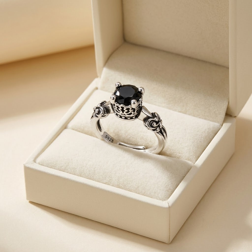 A silver ring with a large round black gemstone and ornate floral details on the band, displayed inside an open cream-colored jewelry box.