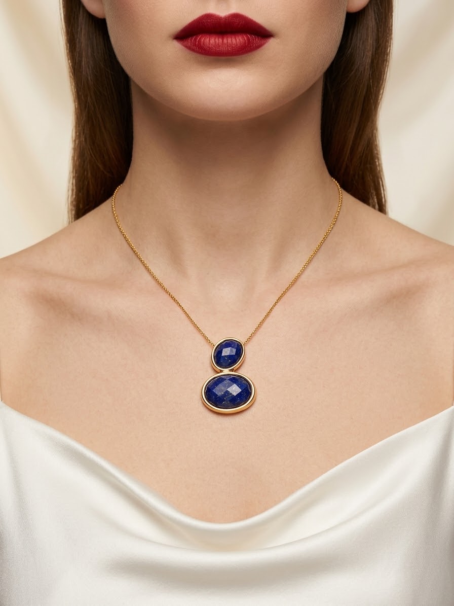 A close-up of a model wearing a delicate gold chain necklace with two stacked oval blue gemstone pendants, styled with a white satin top.