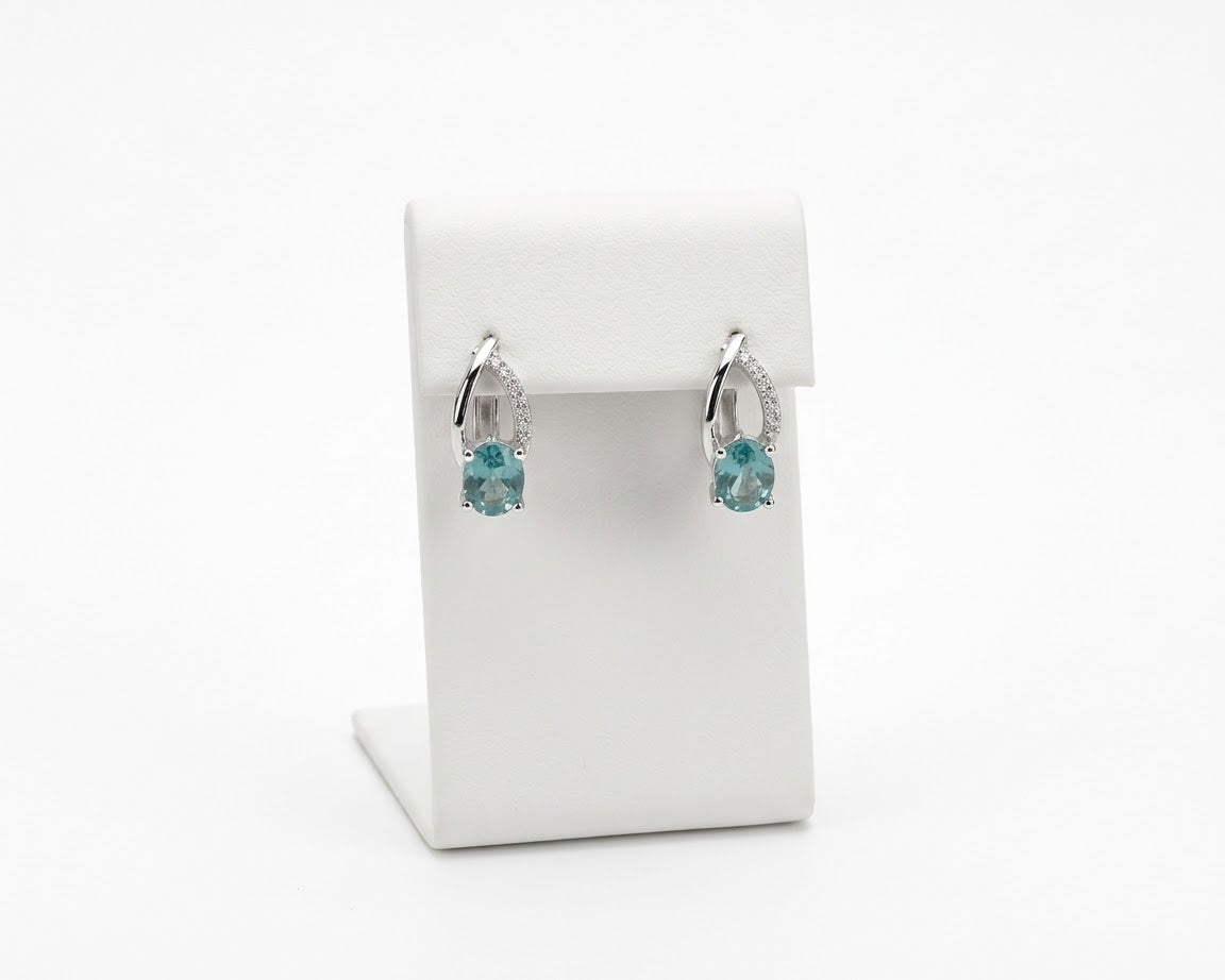 A pair of silver-toned huggie hoop earrings with small clear stones and a central light blue gemstone, displayed on a white jewelry stand against a white background.