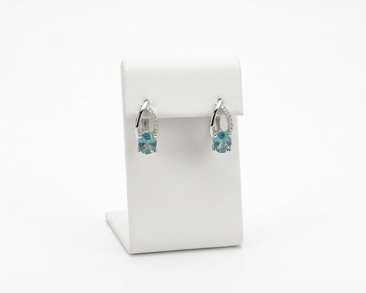 A pair of silver-toned huggie hoop earrings with small clear stones and a central light blue gemstone, displayed on a white jewelry stand against a white background.