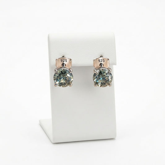 A pair of stud earrings with round iridescent gray-green gemstones in a silver-toned setting, displayed on a white jewelry stand. The back of the earrings is rose gold-toned with a '925' sterling silver mark.