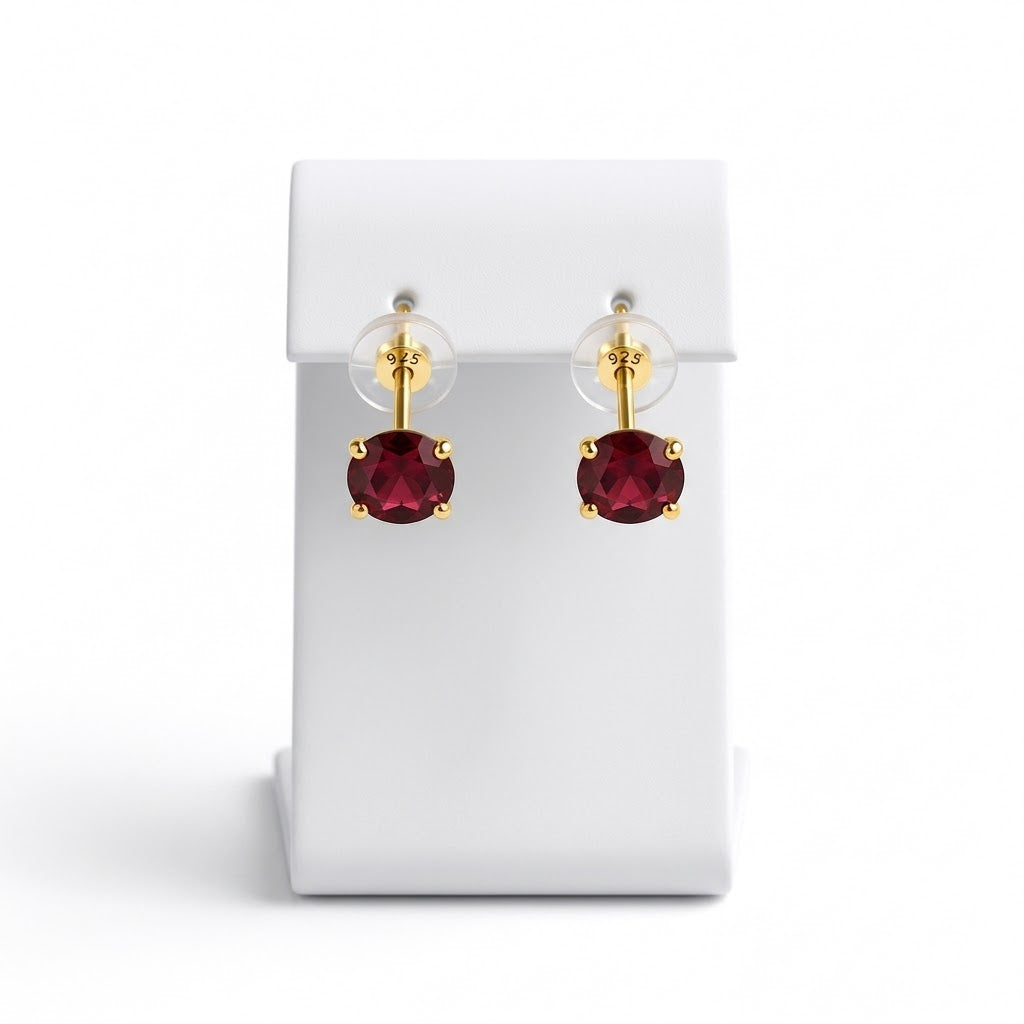 A pair of gold stud earrings with round deep red gemstones, displayed on a white rectangular jewelry stand against a white background.