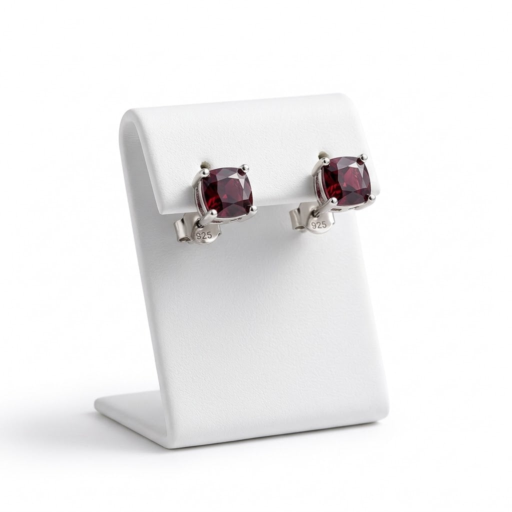 A pair of silver stud earrings with cushion-cut dark red gemstones, displayed on a white jewelry stand against a white background.