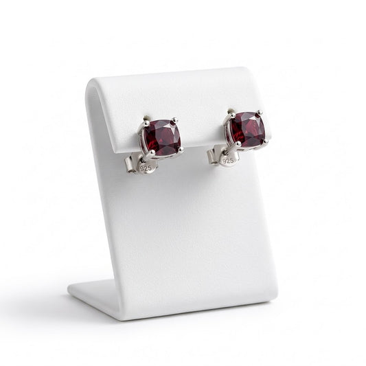 A pair of silver stud earrings with cushion-cut dark red gemstones, displayed on a white jewelry stand against a white background.