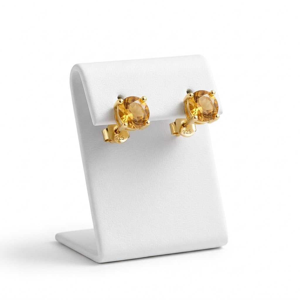 A pair of gold stud earrings with oval-cut yellow gemstones, displayed on a white jewelry stand against a white background.