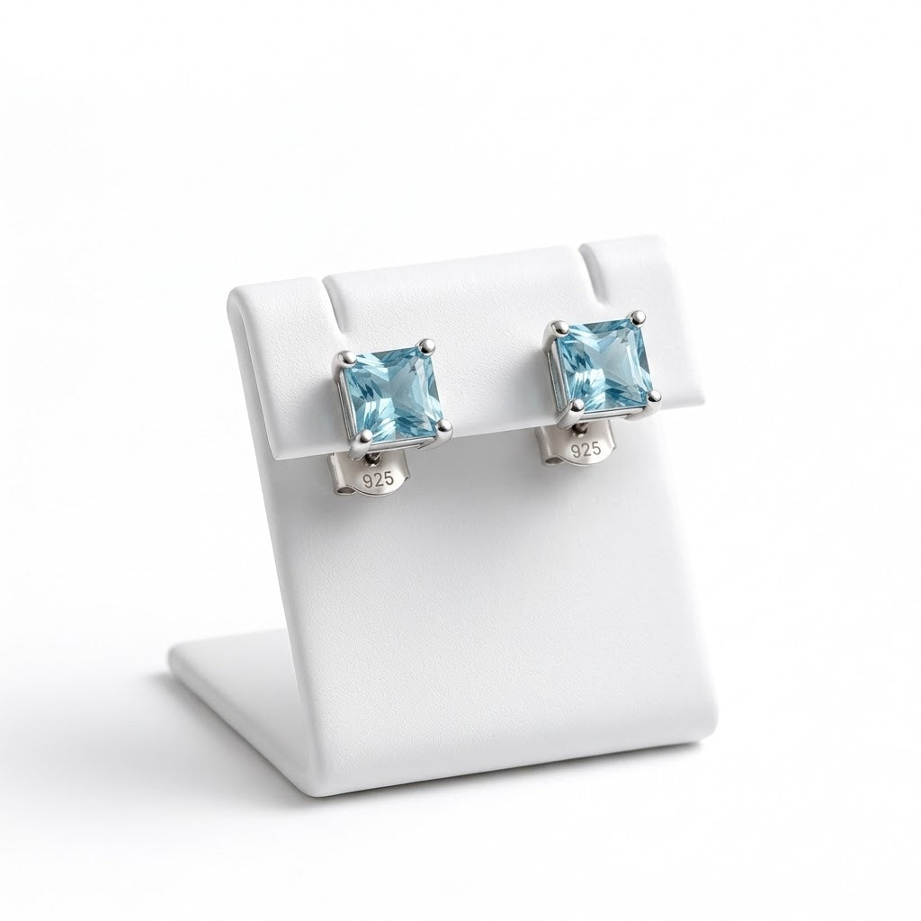 A pair of stud earrings with square light blue gemstones in silver settings, displayed on a white jewelry stand against a white background.