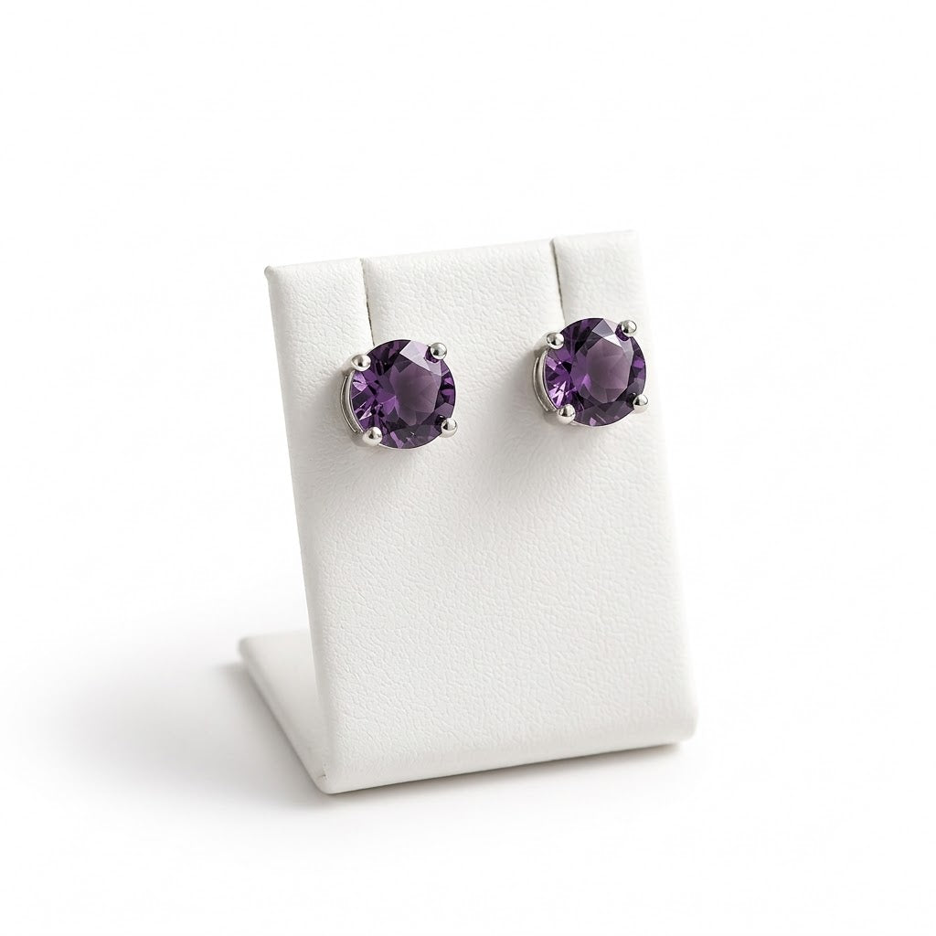 A pair of oval purple gemstone stud earrings in silver prong settings, displayed on a white jewelry stand against a white background.