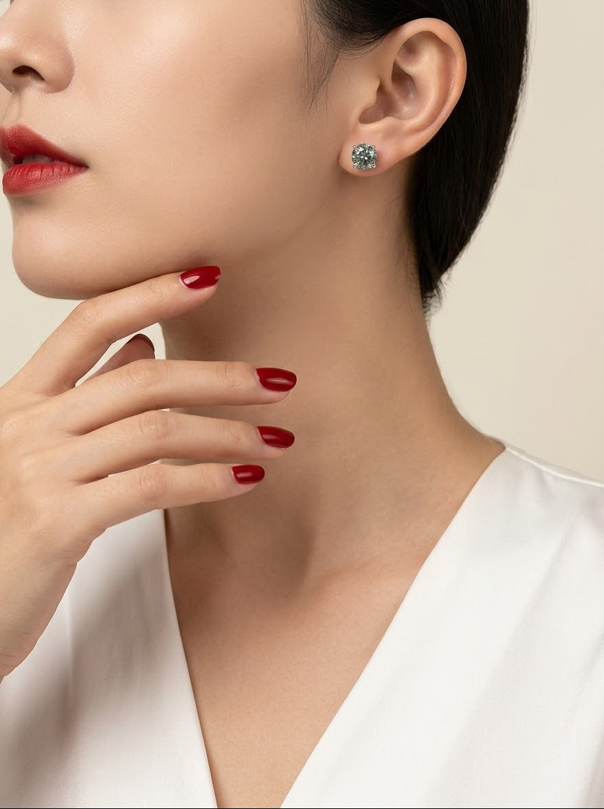 A close-up lifestyle shot of a woman wearing a small round stud earring with a sparkling light blue gemstone. She has dark hair, red lipstick, and red manicured nails, wearing a white top against a soft beige background.