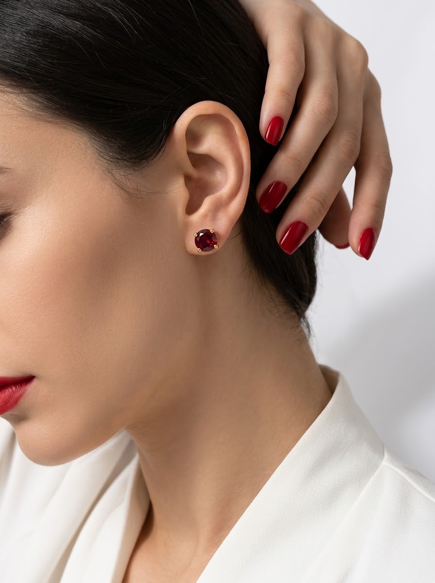 A close-up of a woman's ear wearing a small round red stud earring, with her hand resting near her head and red nail polish visible against a clean white background.