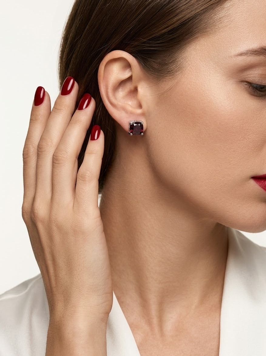 A close-up of a woman wearing a small square stud earring with a dark gemstone in a silver-toned setting, with her hand near her ear against a clean white background.