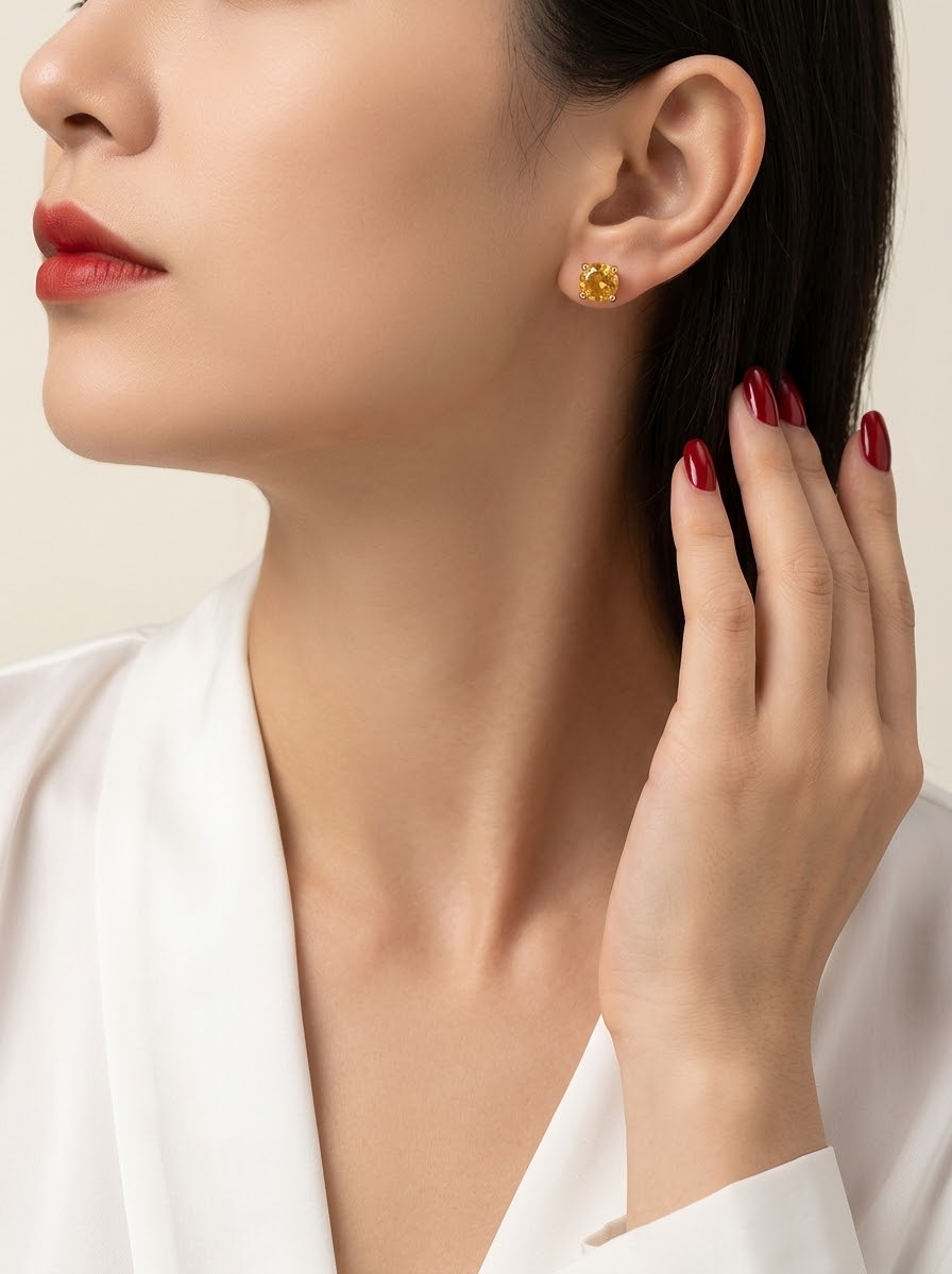 A close-up of a woman wearing a small round yellow gemstone stud earring, with her hand near her hair and a white satin top visible against a soft beige background.